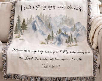 May include: A cream-colored woven throw blanket featuring a watercolor mountain and pine forest landscape. The text reads: I will lift my eyes unto the hills... Where does my help come from? My help comes from the Lord, the maker of heaven and earth. Psalm 121:1-2. The blanket has fringed edges.