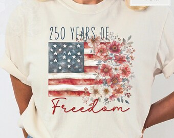 May include: An ivory t-shirt featuring a graphic of the American flag with a watercolor floral arrangement on the right side. The text reads 250 YEARS OF in dark blue and Freedom in red script. The design is centered on the chest of the shirt.