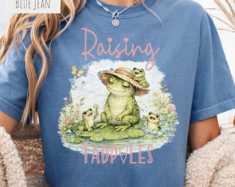 May include: A blue jean colored t-shirt featuring a graphic print of a large frog wearing a straw hat with two smaller frogs on a lily pad. The text "Raising TADPOLES" is printed in pink above and below the illustration with small hearts.