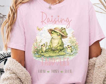 May include: A light pink t-shirt featuring a graphic of a large frog wearing a straw hat with three smaller frogs on a lily pad. The text reads Raising Tadpoles with the names Sarah, James, and Ellie printed below the illustration.