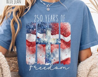 May include: A blue jean colored t-shirt featuring a graphic print with the text 250 YEARS OF Freedom. The design includes a watercolor style American flag background with red, white, and blue colors, and a central cross shape with stars.