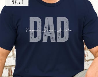 May include: A navy blue short-sleeved t-shirt featuring the word DAD in large, distressed grey block letters. The names Emma, Liam, and Amelia are printed in a thin white script font horizontally across the center of the word DAD.