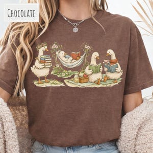 May include: Chocolate-colored t-shirt featuring a whimsical illustration of geese enjoying leisure activities. One goose carries books, another reads in a hammock, and two more read books while enjoying tea. The image includes a picnic basket and floral accents.