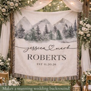 May include: A cream-colored wedding backdrop with a watercolor mountain and forest scene. The names "Jessica & Mark" and "ROBERTS" are printed in black script, with the date "EST 11.20.26" below. The backdrop has a fringed bottom.