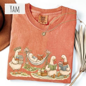 May include: Coral-colored Comfort Colors t-shirt featuring a whimsical illustration of geese and a bear reading books in a park setting. The design includes a bear in a hammock and geese reading books. A gold necklace is draped on the shirt. The word "YAM" is in a white rectangle.