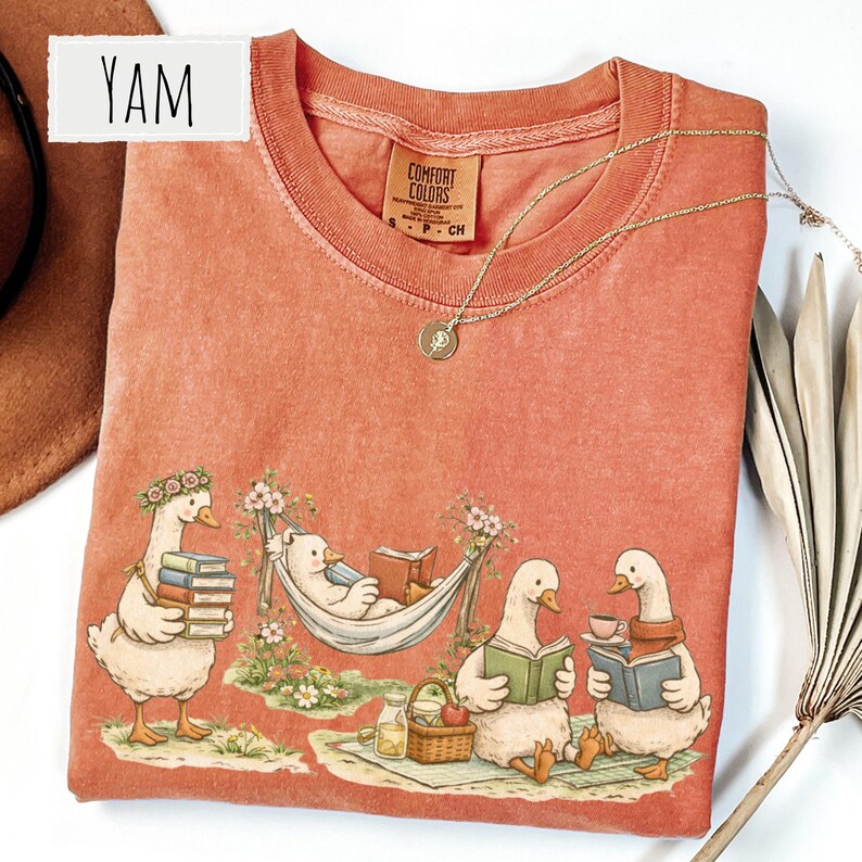 May include: Coral-colored Comfort Colors t-shirt featuring a whimsical illustration of geese and a bear reading books in a park setting. The design includes a bear in a hammock and geese reading books. A gold necklace is draped on the shirt. The word "YAM" is in a white rectangle.