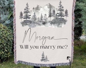May include: A cream-colored woven throw blanket featuring a black and gray watercolor-style mountain and pine tree landscape. The text reads Morgan and Will you marry me? with a small pine tree graphic below. The blanket has a fringed edge.