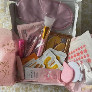 May include: A beauty gift box in pink, containing a sleep mask, lip gloss, face masks, clay masks, a makeup brush, and eye patches. The box is lined with pink tissue paper and has a "Thank You" message. Products are in individual packets.