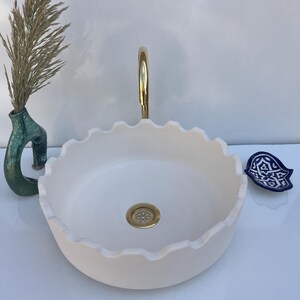 May include: A white, scalloped-edge ceramic sink with a gold tap and drain. A small, blue and white decorative dish sits to the right. A green vase with dried plants is on the left.