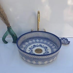 Puede incluir: Un lavabo redondo de cerámica con un patrón geométrico azul y blanco. Cuenta con un grifo de latón y un pequeño jabonera a juego. El diseño presenta detalles intrincados. Un jarrón decorativo con hierba seca está a la izquierda.