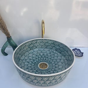 Puede incluir: Un lavabo de baño redondo de cerámica con un patrón de vieiras verde azulado y blanco. El lavabo tiene un grifo y un desagüe dorados. También se ve un pequeño jarrón verde azulado con hierba seca y una decoración de mano de Hamsa azul.