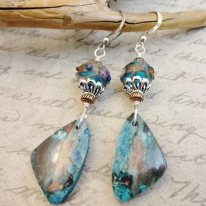 May include: Artisan earrings with a unique design. Each earring features a silver hook, a decorative bead with teal and purple accents, and a teardrop-shaped stone with teal and brown tones. The earrings are displayed against a neutral background.