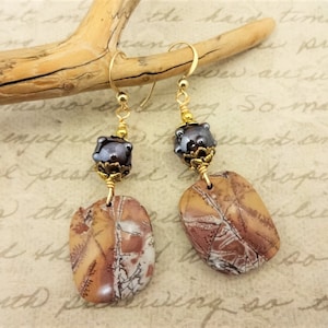 Op de afbeelding: Een paar oorhangers met bruine en witte jaspis stenen, gouden metalen accenten en donkergrijze glazen kralen.