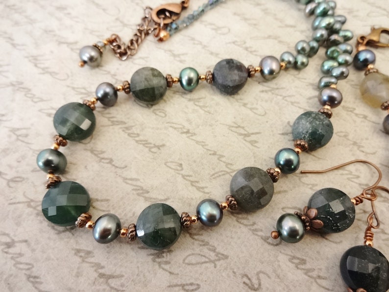 May include: A green and black gemstone necklace and earrings set with copper accents. The necklace features round green and black beads with copper spacers. The earrings are dangle earrings with a green gemstone and copper accents.
