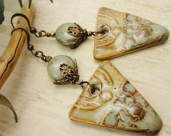 May include: A pair of dangle earrings featuring triangular pendants with a cream, brown, and blue-green pattern. Each earring has a round, light green bead and ornate bronze-colored metal accents. The earrings hang from bronze-colored chains.