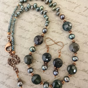 May include: A necklace and earrings set featuring dark green faceted gemstone beads, dark gray pearls, and copper accents. The necklace has a copper clasp and the earrings have infinity-shaped copper hooks.