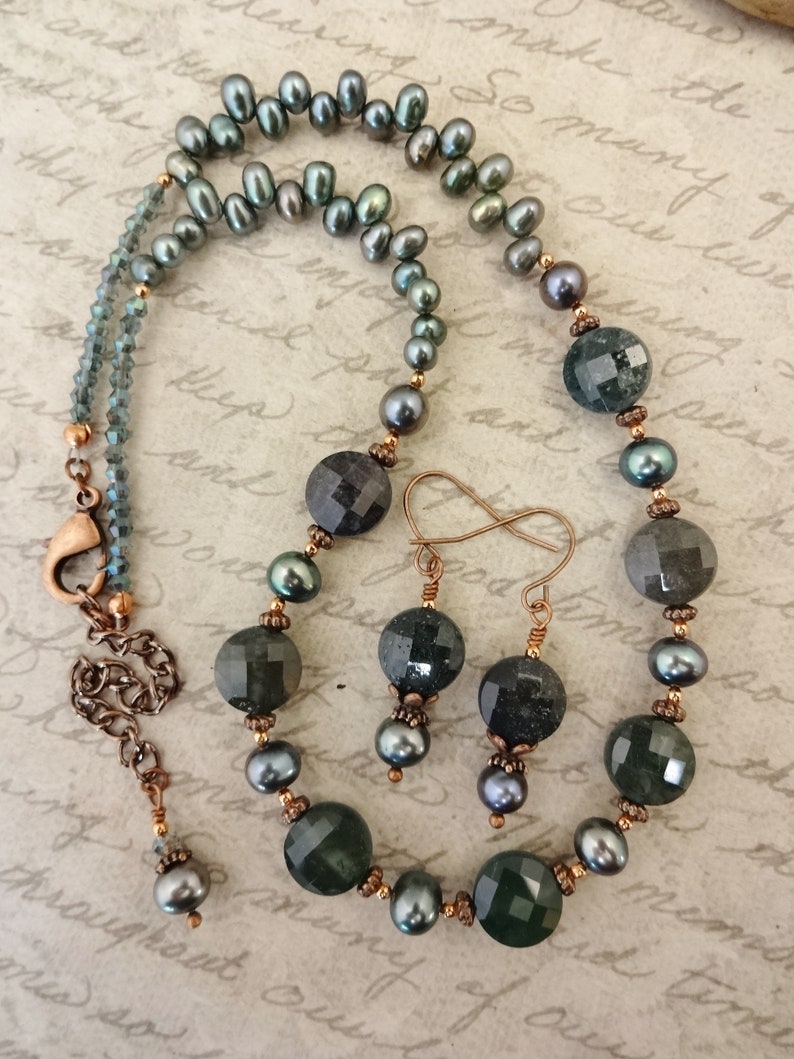 May include: A necklace and earrings set featuring dark green faceted gemstone beads, dark gray pearls, and copper accents. The necklace has a copper clasp and the earrings have infinity-shaped copper hooks.