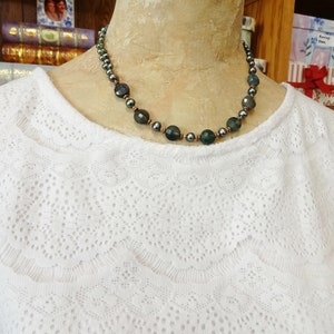 May include: A dark green and black beaded necklace with a copper wire accent. The necklace is on a white lace top.