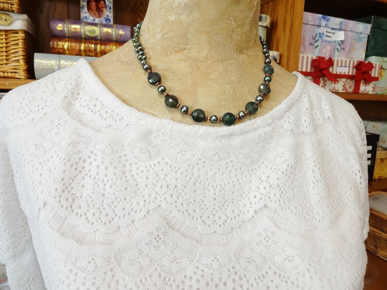 May include: A dark green and black beaded necklace with a copper wire accent. The necklace is on a white lace top.