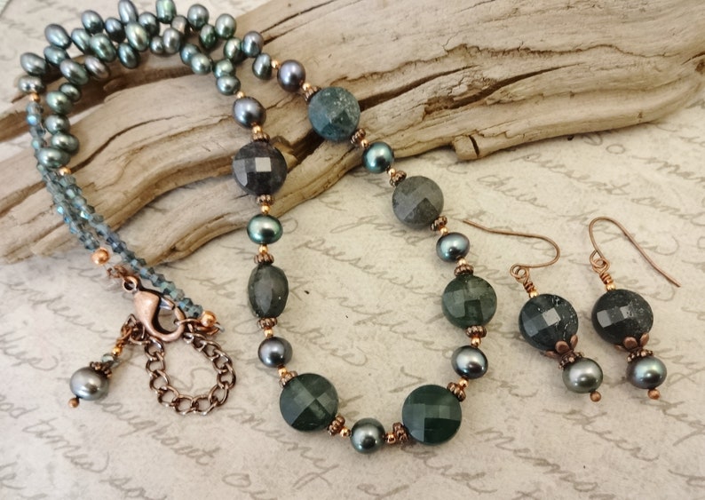 May include: A necklace and earrings set featuring dark green faceted gemstone beads, black pearls, and copper accents. The necklace has a lobster clasp closure.