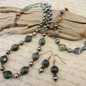 May include: A green and black agate stone necklace and matching earrings with copper accents. The necklace features a layered design with a strand of black pearls and a strand of green agate stones. The earrings are simple and feature a single green agate stone.
