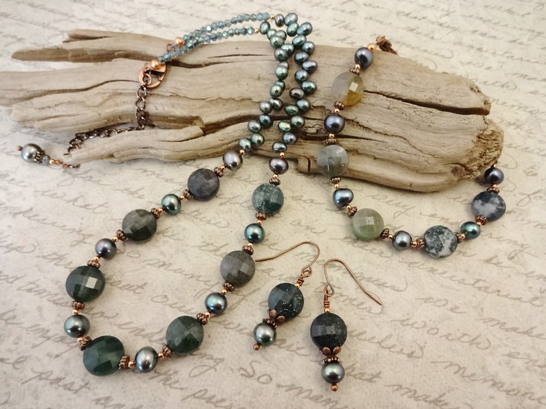 May include: A green and black agate stone necklace and matching earrings with copper accents. The necklace features a layered design with a strand of black pearls and a strand of green agate stones. The earrings are simple and feature a single green agate stone.