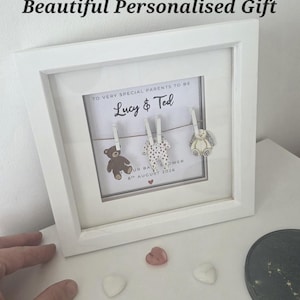 May include: A white square frame containing a personalised print with the text "To very special parents to be Lucy & Ted" and "8th August 2026". Three small wooden clips hold a brown bear, a white polka-dot baby onesie, and a rabbit cutout.