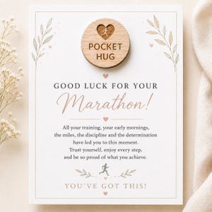 May include: A rectangular card with the text "Good Luck For Your Marathon!" and "You've Got This!" A wooden disc with "Pocket Hug" is attached. The card has a running figure graphic and floral accents.
