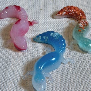 May include: Three small resin lizard figurines. One is translucent pink with glitter, one is a gradient of blue with glitter, and one has a light green body with a textured orange glitter tail featuring small white and orange star-shaped sprinkles.