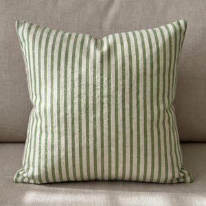 May include: A square scatter cushion featuring a vertical striped pattern in alternating shades of sage green and off-white. The fabric has a visible woven texture, and the cushion is filled to maintain a plump, structured square shape.