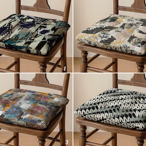 May include: Four wooden chairs each with a different patterned cushion. The cushions feature abstract designs in various colors, including blue, green, yellow, black, and white. The chairs are made of wood and have a classic design.