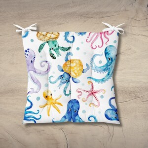 May include: A square seat cushion featuring a white background with a colorful watercolor pattern of sea creatures including octopuses in purple, blue, and yellow, a sea turtle, and a pink starfish, with white fabric ties at the top corners.