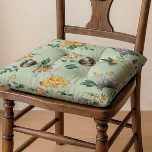 Könnte beinhalten: Ein quadratisches Kissen mit Blumenmuster liegt auf einem Holzstuhl. Das Kissen zeigt gelbe Rosen, graue und braune Blumen und grüne Blätter auf hellgrünem Hintergrund. Der Stuhl ist aus dunklem Holz gefertigt.