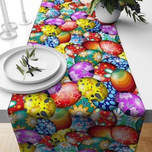 May include: A colorful Easter-themed table runner featuring a pattern of decorated Easter eggs in various colors, including yellow, purple, red, and blue. The eggs have floral and star designs. The runner is placed on a white table.