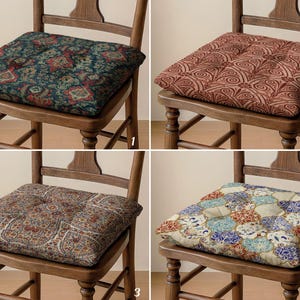 Könnte beinhalten: Vier quadratische Stuhlkissen mit unterschiedlichen Mustern. Die Kissen sind auf Holzstühlen platziert. Das erste Kissen hat einen dunkelblauen Hintergrund mit roten und goldenen Blumenmustern. Das zweite Kissen hat ein rotes und braunes Wirbelmuster. Das dritte Kissen hat ein braunes Paisley-Muster. Das vierte Kissen hat ein blaues, rotes und goldenes Blumenmuster.