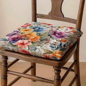 以下が含まれることがあります： 赤、オレンジ、黄色、紫、青の色合いの花柄の正方形のクッションが木製の椅子の上にあります。クッションのデザインは、白い背景に対して水彩画風の花と葉の配置が特徴です。