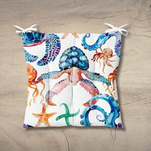 May include: Square cushion featuring a watercolor design of sea creatures. The cushion showcases a central sea turtle surrounded by starfish, jellyfish, and octopus in shades of blue, orange, and green on a white background. White ties are attached to the corners.