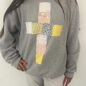 May include: A grey sweatshirt featuring a patchwork cross design. The cross is made from fabric squares in floral and patterned prints, including pink, yellow, and blue. The sweatshirt is a casual, comfortable style.