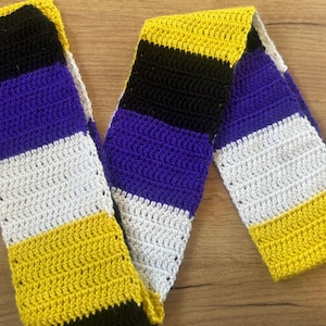 May include: A long, rectangular crochet scarf featuring horizontal stripes in alternating colours of black, purple, white, and yellow. The textile is crafted with a consistent double crochet stitch pattern throughout the entire length of the accessory.