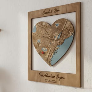 Peut inclure: Carte en forme de cœur en bois avec un plan de ville, encadrée dans un cadre rectangulaire en bois. La carte présente une zone d'eau bleue et une épingle en forme de cœur rouge. Le cadre comprend le texte "Sarah & Ben" et "Our Adventure Begins 07-15-2025".