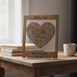 Può includere: Una mappa a forma di cuore incorniciata in legno con un fronte in acrilico trasparente. La mappa presenta un layout della città con elementi d'acqua blu e un indicatore rosso. La cornice riporta i nomi "David & Sarah" e il testo "Where Our Story Began 08-15-2024".