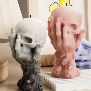 May include: Two skull-shaped candles. One is white and grey, the other is pink. Both candles have a hand-like structure holding the skull. The base of each candle is decorated with small skulls. The candles are on a white surface.