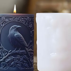 Puede incluir: Una vela azul oscuro encendida con un diseño detallado de cuervo en un marco circular, rodeado de elementos florales. La vela está junto a una vela blanca sin encender. El cuervo está posado en una rama.