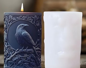 Può includere: Una candela blu scuro accesa con un design dettagliato di corvo in una cornice circolare, circondata da elementi floreali. La candela è accanto a una candela bianca non accesa. Il corvo è appollaiato su un ramo.