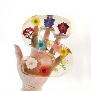 May include: A clear resin letter "S" filled with colorful dried flowers. The flowers include yellow daisies, blue delphiniums, and pink blossoms. The resin has a gold glitter effect, and the letter is held in a hand.