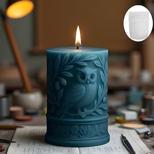 Pode incluir: Uma vela de pilar azul-petróleo com um desenho de coruja e ramo de oliveira. A vela está acesa, com uma chama no topo. A coruja está no centro da vela, rodeada por entalhes detalhados. A vela está numa secretária.