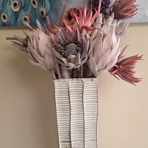 May include: A tall, rectangular white vase with a textured, horizontal stripe design, filled with dried flowers in shades of pink, purple, and beige. The vase sits on a small, round table with a marble top. A painting of a peacock is in the background.