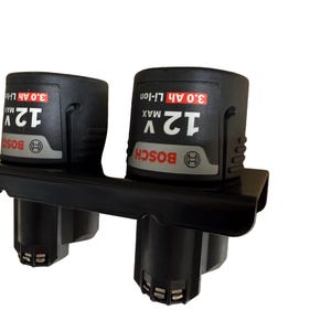May include: A black plastic wall mount holder containing two Bosch 12V Max 3.0 Ah Li-Ion batteries. The batteries are black with red and white labels featuring the Bosch logo, voltage, and capacity specifications clearly printed on the front.