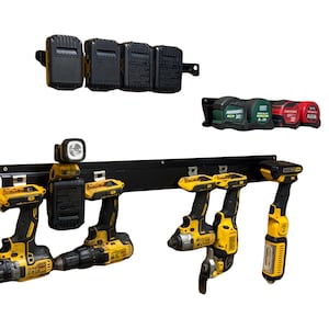 May include: A black metal wall-mounted tool storage system featuring a long horizontal rail holding four yellow and black power tools, a battery, and a work light. Above, a separate battery rack holds four batteries and a tape measure rack holds four tapes.