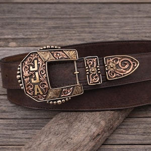 Puede incluir: Un cinturón de cuero marrón con una hebilla decorativa. La hebilla es dorada y cobriza, con las iniciales "JJA" y adornos ornamentados. El cinturón está sobre un fondo de madera rústica.